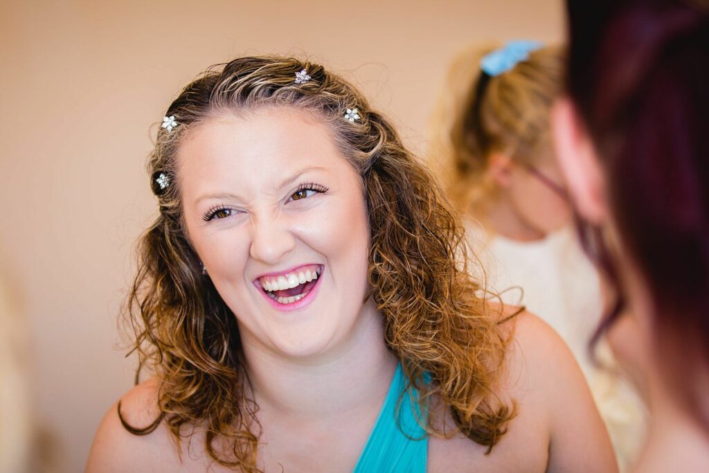 A woman with curly hair, adorned with small floral decorations, wearing a blue sleeveless top, is laughing joyfully. The background is softly blurred with another person slightly visible. The overall atmosphere appears to be cheerful and lively. Image by Documentary Wedding Photographer Lewis Bishop Photography.