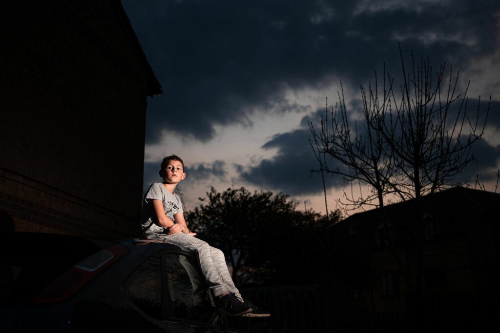 A young boy sits on the roof of a car at dusk, illuminated by a soft light, with dark clouds and silhouettes of trees and buildings in the background. He gazes pensively into the distance, wearing a light-colored outfit and sneakers. Image by Documentary Wedding Photographer Lewis Bishop Photography.