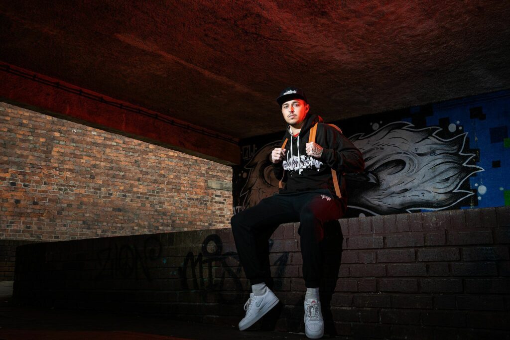 A person wearing dark streetwear, a cap, and white sneakers sits on a brick ledge in an urban area. They have a backpack on and face the camera in an underpass with graffiti-covered walls illuminated by red and white lights. Image by Documentary Wedding Photographer Lewis Bishop Photography.