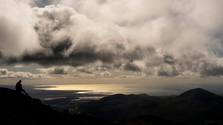 A lone person sits on a rocky hillside under thick, dramatic clouds. The sun shines through breaks in the clouds, casting light over a distant body of water and the landscape below. Rolling hills extend towards the horizon. Image by Documentary Wedding Photographer Lewis Bishop Photography.