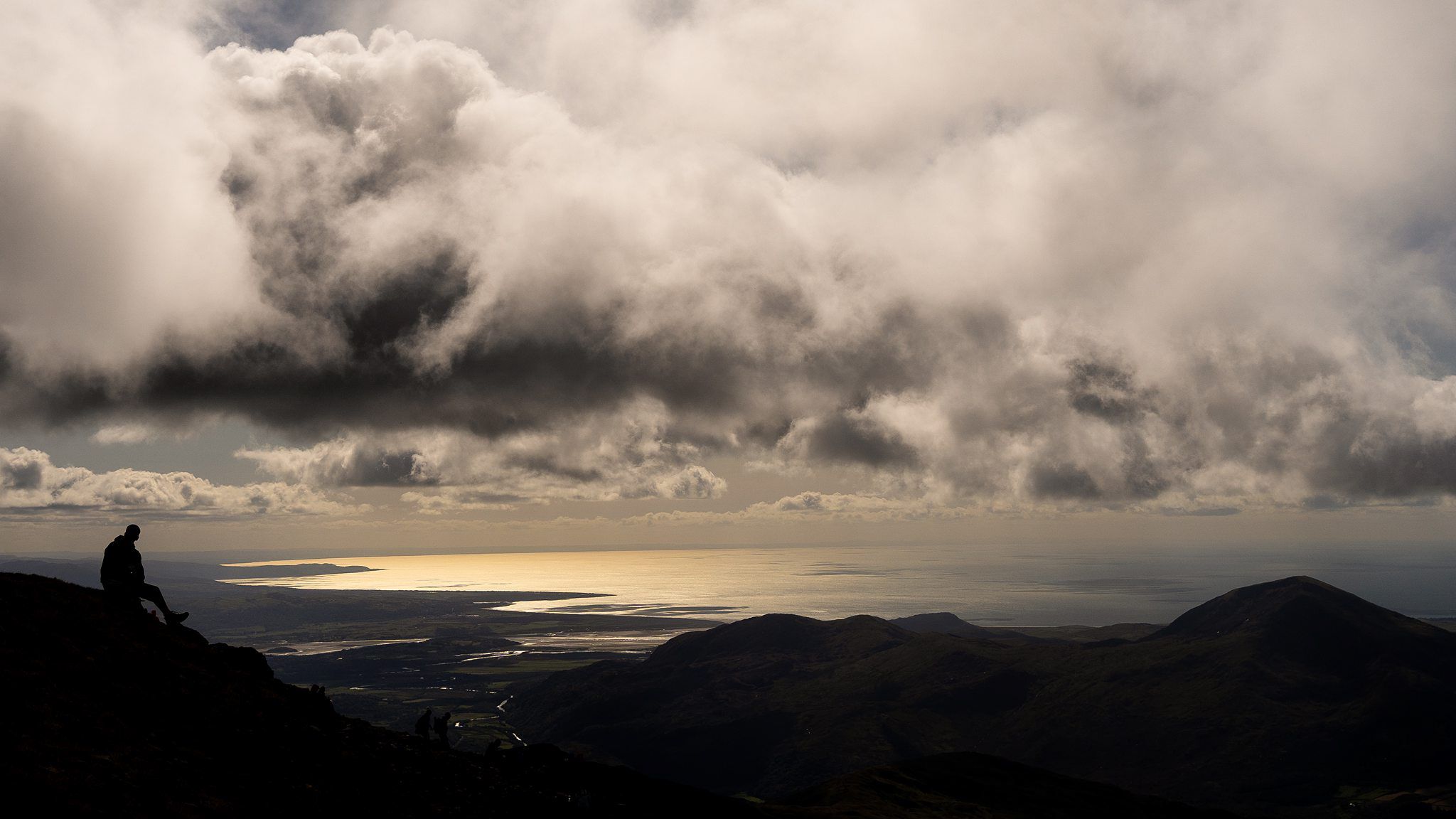 A lone person sits on a rocky hillside under thick, dramatic clouds. The sun shines through breaks in the clouds, casting light over a distant body of water and the landscape below. Rolling hills extend towards the horizon. Image by Documentary Wedding Photographer Lewis Bishop Photography.