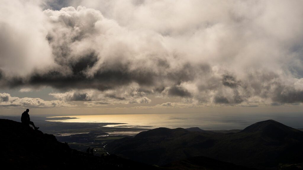 A lone hiker sits on a mountain peak, looking out over a vast landscape with rolling hills and a shimmering body of water illuminated by sunlight breaking through the clouds. The sky is filled with dramatic, heavy clouds. Image by Documentary Wedding Photographer Lewis Bishop Photography.