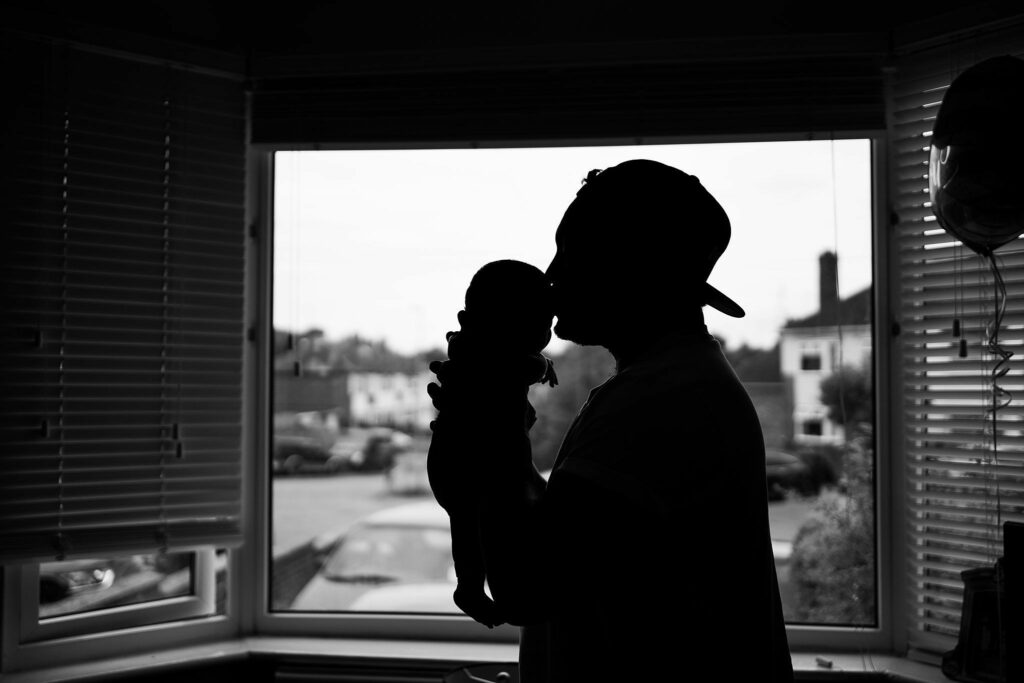 Silhouette of a person in a cap gently kissing an infant inside a room with large windows. A residential neighborhood is visible through the window, partially obscured by partially closed blinds. Image by Documentary Wedding Photographer Lewis Bishop Photography.