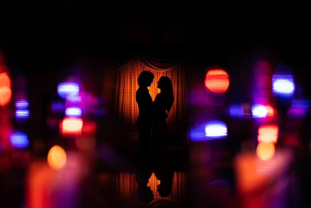 A silhouette of a couple stands close together, facing each other against a warm, orangish backdrop. The scene is accentuated by out-of-focus, colorful lights in the foreground, creating a soft, romantic ambiance. Image by Documentary Wedding Photographer Lewis Bishop Photography.