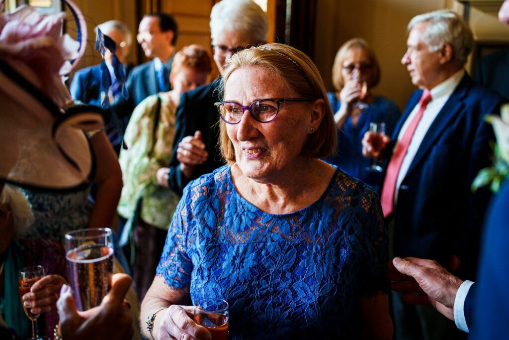 An elderly woman with short blonde hair and glasses, wearing a blue lace dress, is engaged in conversation at a social gathering. She holds a drink in her hand. Several people, dressed in formal attire, are conversing and mingling in the background. Image by Documentary Wedding Photographer Lewis Bishop Photography.