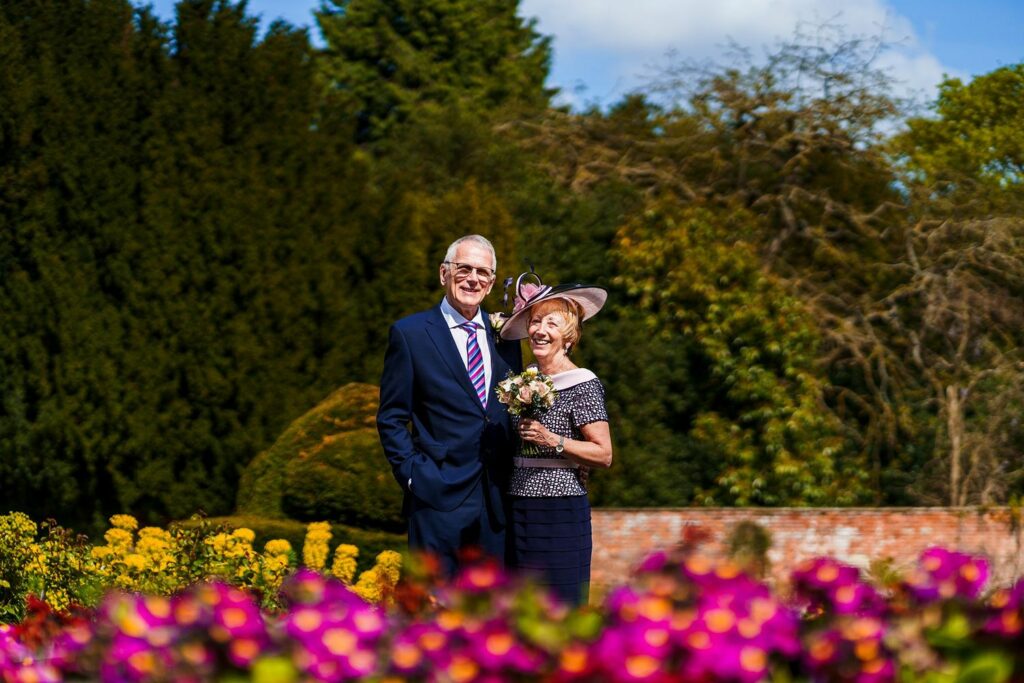 An elderly couple stands smiling outdoors in a garden filled with vibrant flowers. The man is in a suit and tie, while the woman wears a dress and hat, holding a small flower bouquet.  Tall trees and a brick wall form the background under a partly cloudy sky. Image by Documentary Wedding Photographer Lewis Bishop Photography.