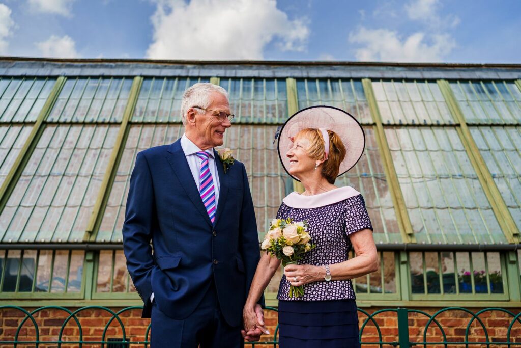 An elderly couple dressed in formal attire stands hand in hand, smiling at each other. The man is in a blue suit with a striped tie, and the woman is holding a bouquet, wearing a hat and a patterned dress. They are in front of a greenhouse with a blue sky above. Image by Documentary Wedding Photographer Lewis Bishop Photography.
