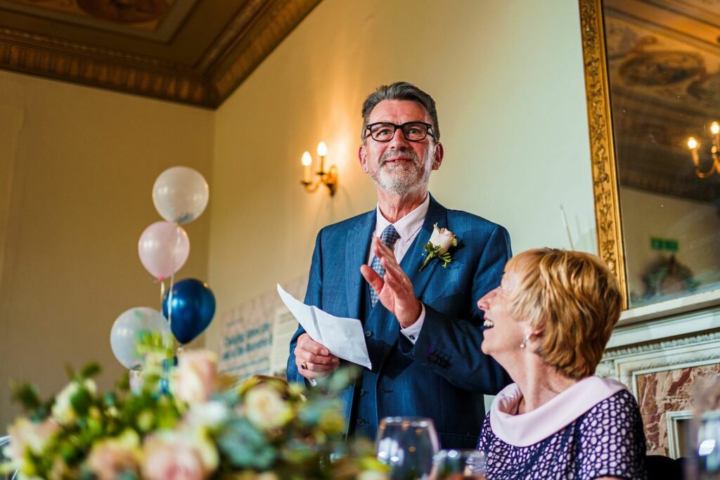 A man in a suit and glasses is speaking enthusiastically and holding a piece of paper while a woman beside him smiles. They are in a decorated room with flowers, balloons, and a chandelier, suggesting a formal event or celebration. Image by Documentary Wedding Photographer Lewis Bishop Photography.