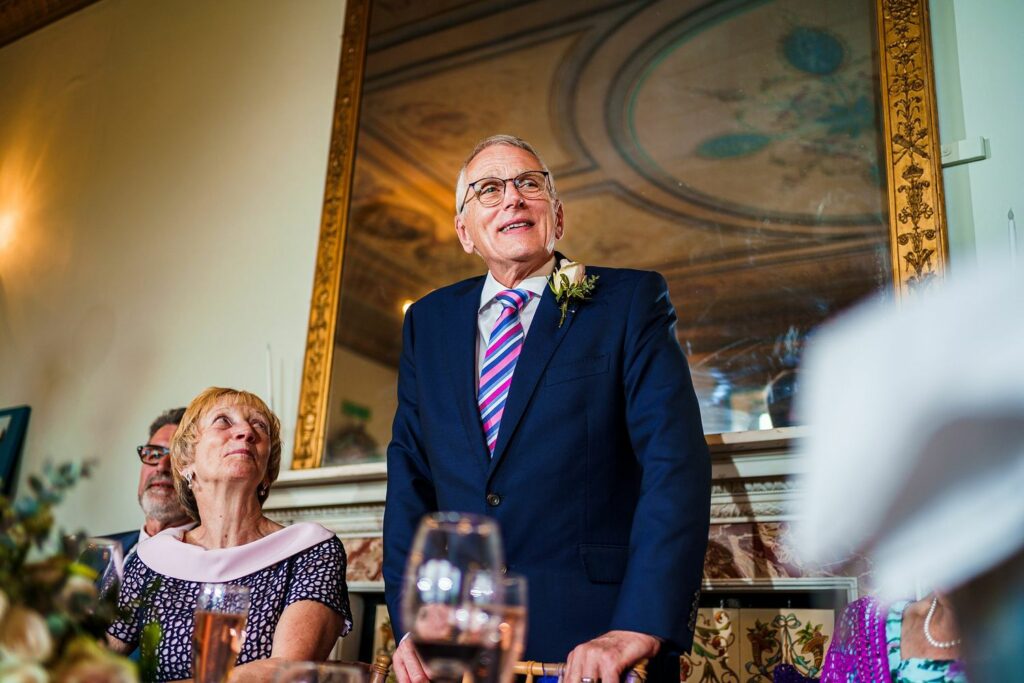 An older man in a suit and striped tie stands and addresses a group at a formal event. A woman seated next to him looks up at him attentively. The room is elegantly decorated with a large antique mirror and ornate ceiling. Glasses and floral arrangements are on the table. Image by Documentary Wedding Photographer Lewis Bishop Photography.