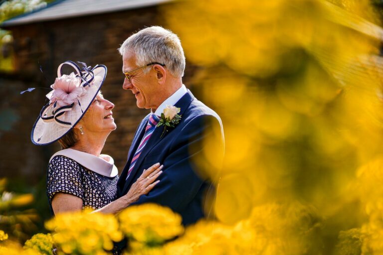 An elderly couple gazes lovingly into each other's eyes, standing close together amid vibrant yellow flowers. The woman wears a decorative hat with flowers and netting; the man wears glasses, a suit, and a striped tie. A blurred background adds a warm, romantic ambiance. Image by Documentary Wedding Photographer Lewis Bishop Photography.