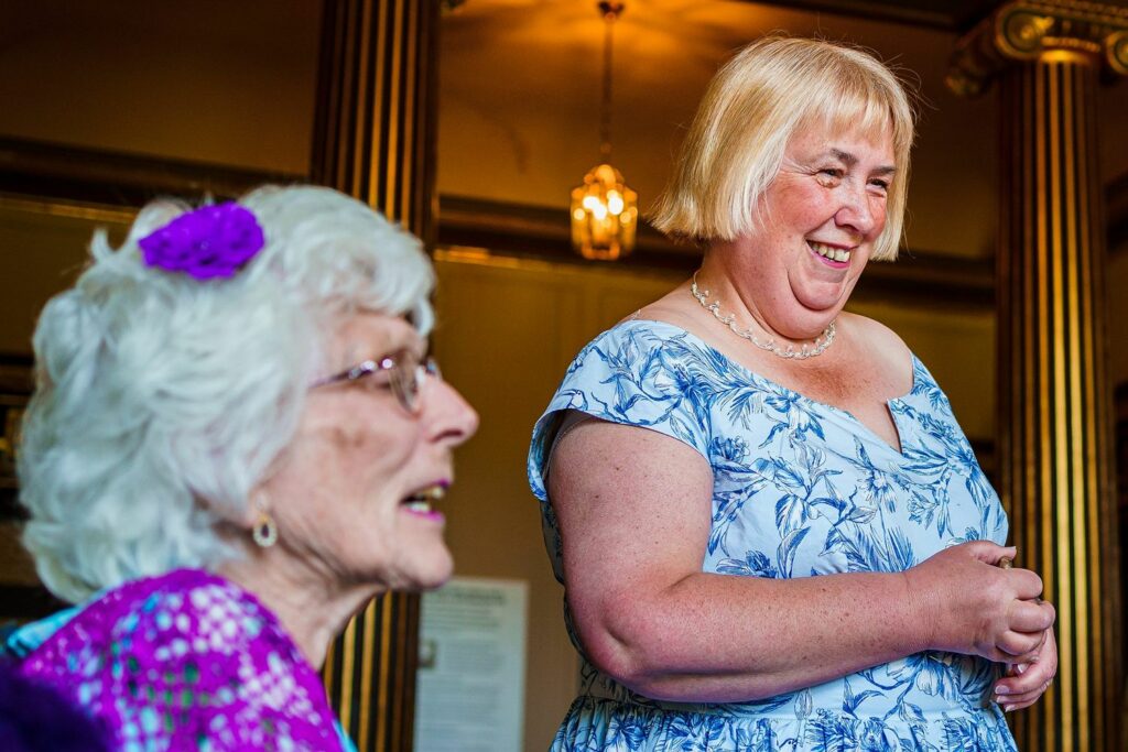 An elderly woman with white hair and glasses, adorned with a purple flower in her hair, smiles while sitting in the foreground. A blonde woman in a blue floral dress stands smiling and looking at her, against a backdrop of decorative gold columns and warm lighting. Image by Documentary Wedding Photographer Lewis Bishop Photography.