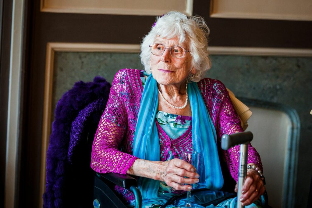 Elderly person with short white hair sits in a wheelchair, dressed in vibrant colorful clothes. They hold a drink in one hand and appear thoughtful. The background shows a warmly lit indoor setting with a mix of light and dark tones. Image by Documentary Wedding Photographer Lewis Bishop Photography.