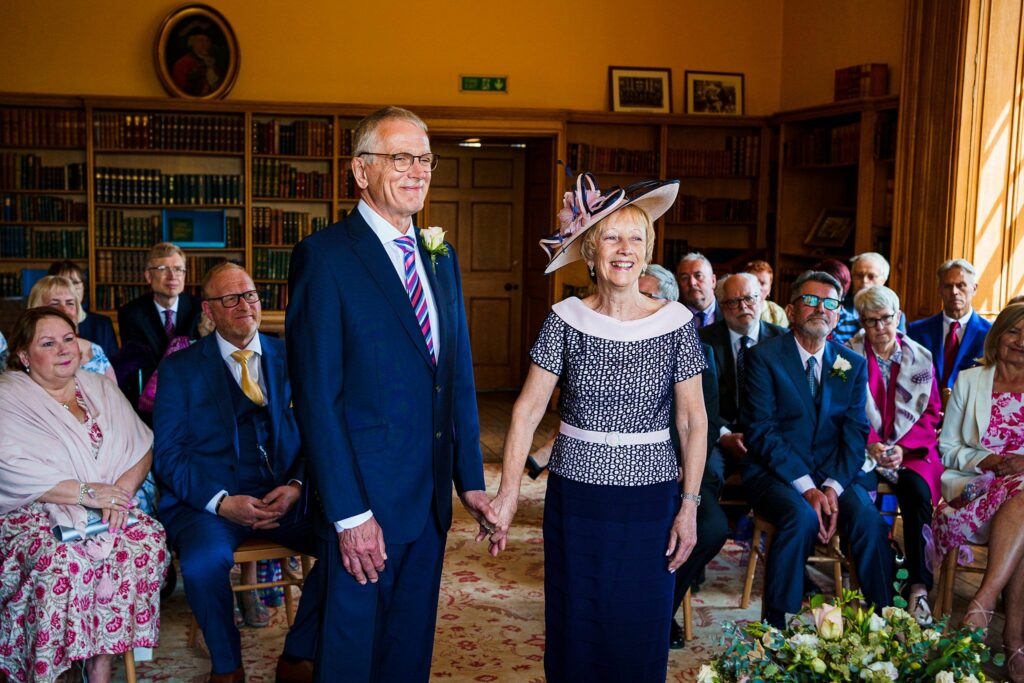 A senior couple stands holding hands in a well-lit library room, surrounded by seated friends and family. The man wears a navy suit and striped tie, and the woman is dressed in a navy dress with a patterned top and a large hat. Everyone is smiling. Image by Documentary Wedding Photographer Lewis Bishop Photography.