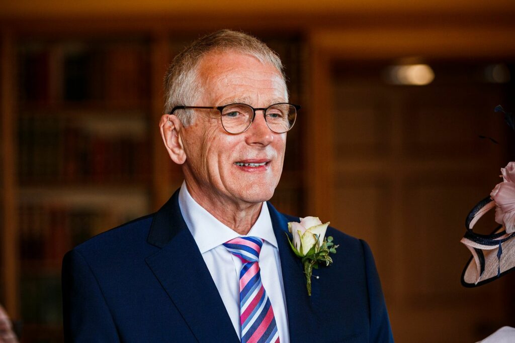 An older man with short gray hair and glasses is wearing a dark blue suit, a striped tie, and a boutonnière with a white rose and greenery. He appears to be in a warmly lit room with bookshelves in the background. Image by Documentary Wedding Photographer Lewis Bishop Photography.