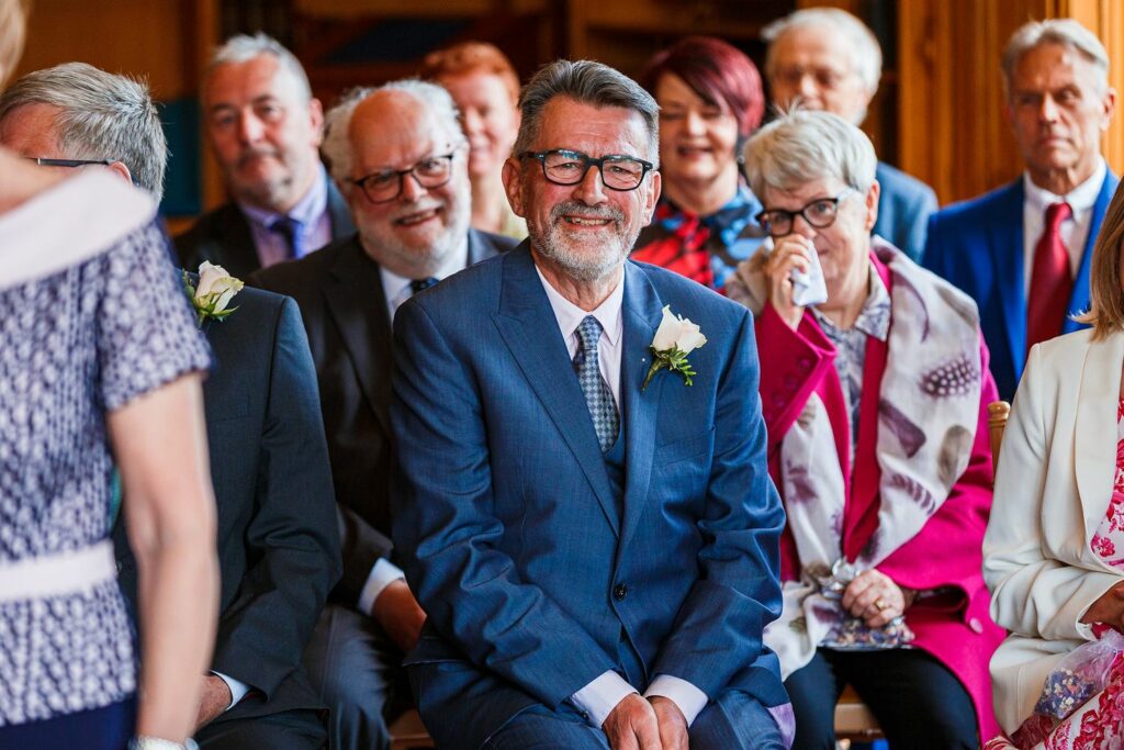 A smiling man in a blue suit, wearing glasses and a boutonniere, is seated in a decorated indoor setting with people dressed in formal attire sitting around him. The atmosphere is joyful, with a woman in the background dabbing her eyes with a tissue. Image by Documentary Wedding Photographer Lewis Bishop Photography.