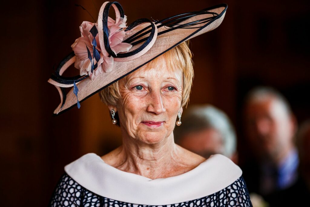 An elderly woman is dressed elegantly in a black and white outfit, featuring a distinctive wide-brimmed hat adorned with pink flowers and black loops. She is indoors, with blurred figures in the background. Her expression is calm and serene. Image by Documentary Wedding Photographer Lewis Bishop Photography.