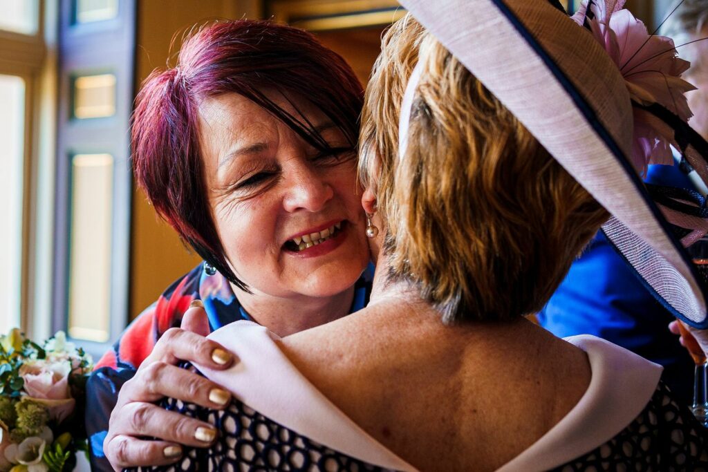 Two women embrace happily, one with short dark hair smiling warmly, the other with lighter hair wearing a hat adorned with flowers. Bright natural light from a nearby window illuminates the scene, reflecting a cheerful and warm atmosphere. Image by Documentary Wedding Photographer Lewis Bishop Photography.