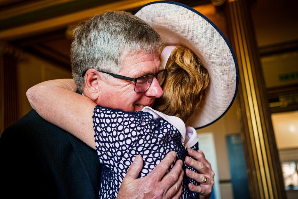 A man with gray hair and glasses warmly embraces a woman wearing a large, light-colored hat with a pink ribbon. Both are smiling and dressed in formal attire, surrounded by ornate decor with golden pillars in the background. Image by Documentary Wedding Photographer Lewis Bishop Photography.