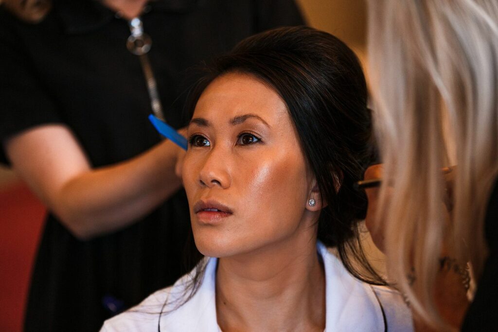 A woman with dark hair is getting her makeup done by two stylists. One stylist is applying makeup near her eyes, while the other is working on her hair. She is wearing a white top and looking off into the distance. The background is blurred. Image by Documentary Wedding Photographer Lewis Bishop Photography.