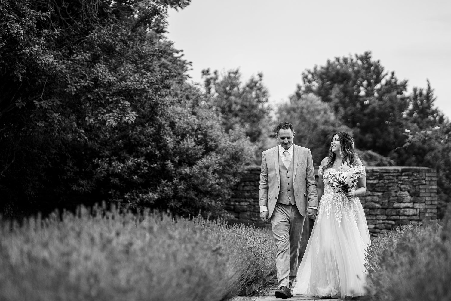A bride and groom walk hand in hand in an outdoor garden setting. The bride wears a white gown and holds a bouquet; the groom is in a light-colored suit. Image by Documentary Wedding Photographer Lewis Bishop Photography.