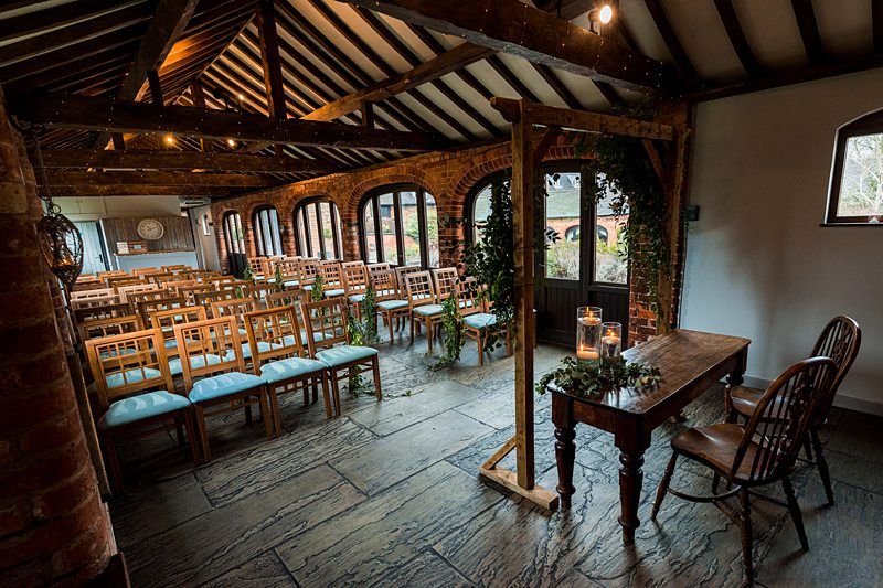 A rustic indoor wedding venue with wooden beams, stone floors, and rows of wooden chairs with blue cushions. There's a wooden table with two chairs near a large window, and greenery decorates the space. Candles are placed on the table, creating a cozy ambiance. Image by Documentary Wedding Photographer Lewis Bishop Photography.