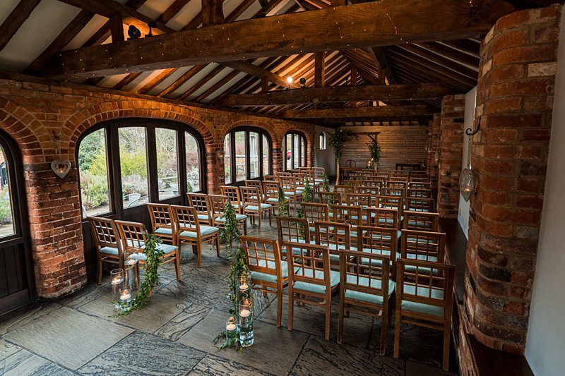 A rustic indoor venue with brick walls and wooden beams is set up for a ceremony. Wooden chairs with green cushions are arranged in rows. Greenery and lanterns line the aisle, and large windows allow natural light to fill the space. Image by Documentary Wedding Photographer Lewis Bishop Photography.