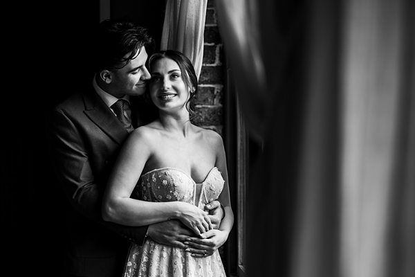 Black and white photo of a couple standing by a window with curtains. The man in a suit is behind the woman, embracing her from behind. The woman is wearing a strapless dress and smiling, looking out the window while the man gazes at her lovingly. Image by Documentary Wedding Photographer Lewis Bishop Photography.
