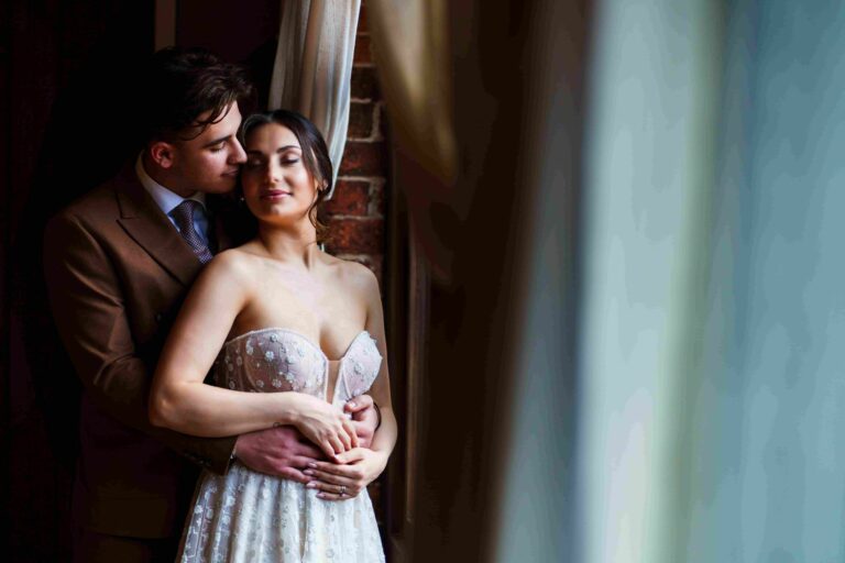 A couple stands closely by a window with cream-colored curtains. The man, dressed in a brown suit, embraces the woman from behind, resting his chin near her ear. The woman, wearing a strapless white dress with floral details, rests her hands on his and closes her eyes. Image by Documentary Wedding Photographer Lewis Bishop Photography.