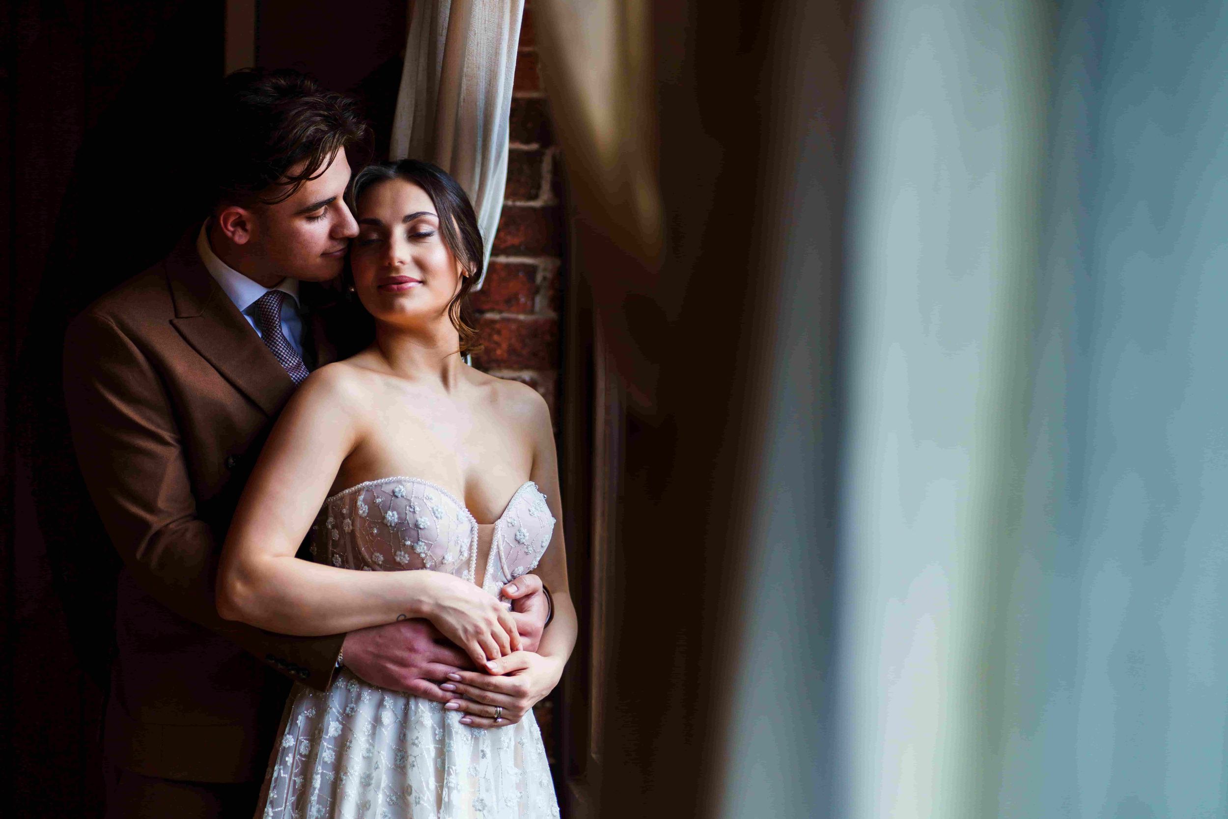 A couple stands closely by a window with cream-colored curtains. The man, dressed in a brown suit, embraces the woman from behind, resting his chin near her ear. The woman, wearing a strapless white dress with floral details, rests her hands on his and closes her eyes. Image by Documentary Wedding Photographer Lewis Bishop Photography.
