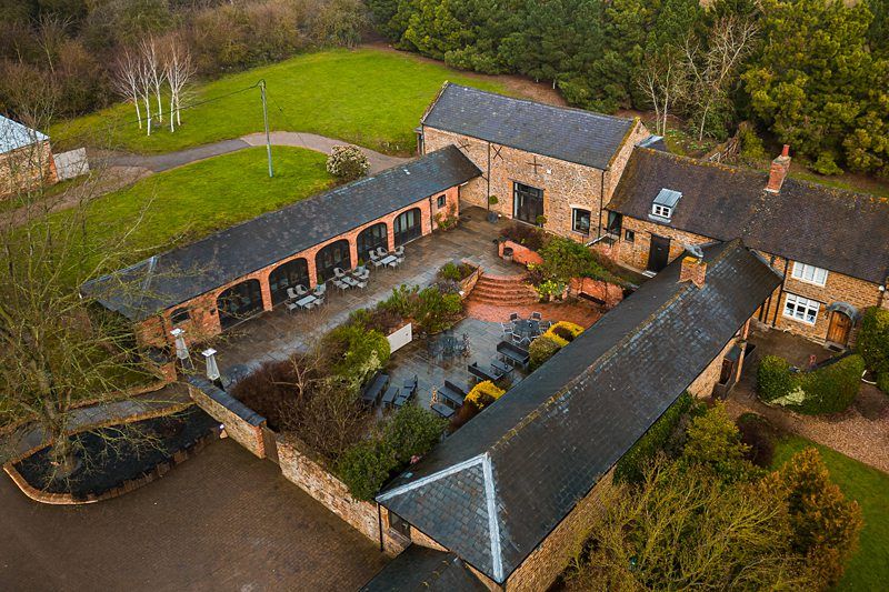 Aerial view of a rustic courtyard surrounded by brick buildings with slate roofs. There are outdoor seating areas with tables and chairs, greenery, and a cobblestone pathway. Tree-lined grassy areas are seen in the background. Image by Documentary Wedding Photographer Lewis Bishop Photography.