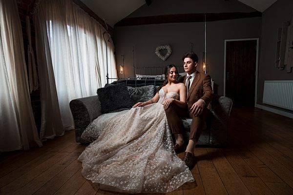 A bride and groom sit on a sofa in a dimly lit room with wooden floors and a rustic decor. The bride wears a flowing, white dress with lace detailing, and the groom is dressed in a brown suit. They pose together, the bride leaning on the groom, with curtains in the background. Image by Documentary Wedding Photographer Lewis Bishop Photography.
