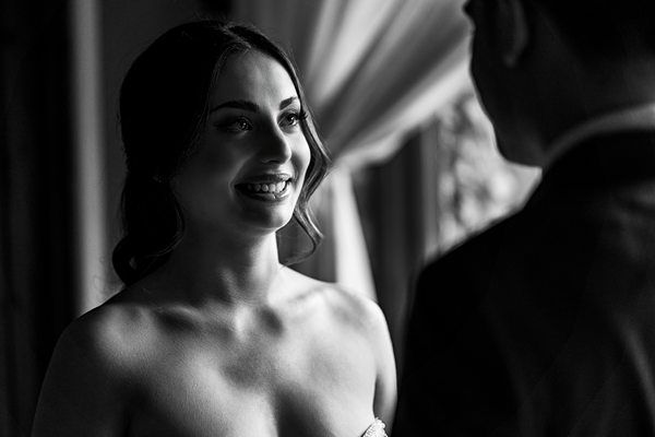 A black and white photo of a young woman smiling warmly at a man, who is turned away from the camera. The woman has long hair tied back and appears to be wearing an elegant dress. The background is softly lit with curtains and a hint of a window. Image by Documentary Wedding Photographer Lewis Bishop Photography.