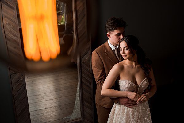 A couple dressed in elegant wedding attire share an intimate moment. The man in a brown suit embraces the woman from behind, who is wearing a white, strapless gown. They stand near a wooden wall with a warm, glowing light out of focus in the foreground. Image by Documentary Wedding Photographer Lewis Bishop Photography.