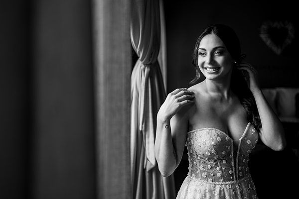 A black and white photo of a smiling bride with long wavy hair in an embroidered strapless gown. She stands near a window with a curtain tied back, touching her hair and looking out with a joyful expression. A heart-shaped decor is visible on the wall behind her. Image by Documentary Wedding Photographer Lewis Bishop Photography.