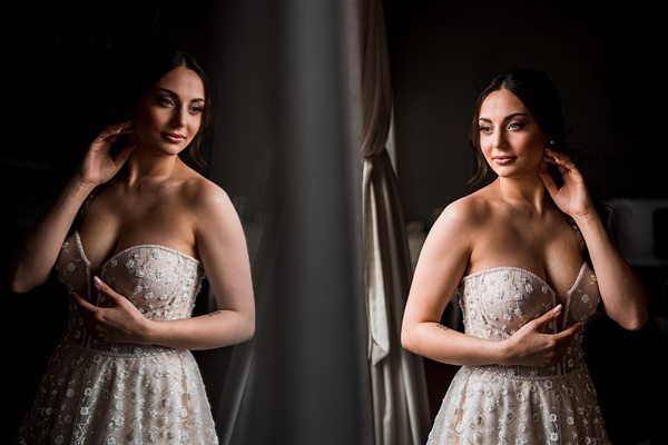 A woman wearing a strapless, detailed dress gazes thoughtfully into the distance, while posing beside a reflective surface that mirrors her image. The lighting accentuates her features and the intricate details of her dress. Image by Documentary Wedding Photographer Lewis Bishop Photography.