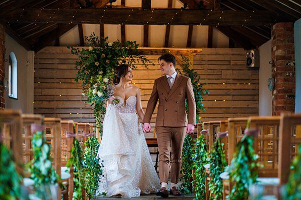 A bride in a white wedding dress and a groom in a brown suit walk hand-in-hand down an aisle decorated with greenery inside a rustic venue. They are smiling at each other, surrounded by wooden chairs and fairy lights hanging from the wooden beams above. Image by Documentary Wedding Photographer Lewis Bishop Photography.
