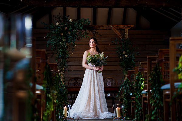 A bride in a white, flowing wedding dress stands at the end of a decorated aisle in a rustic venue. She holds a bouquet of flowers and smiles, with candles and greenery adorning the aisle and a wooden backdrop behind her. Image by Documentary Wedding Photographer Lewis Bishop Photography.