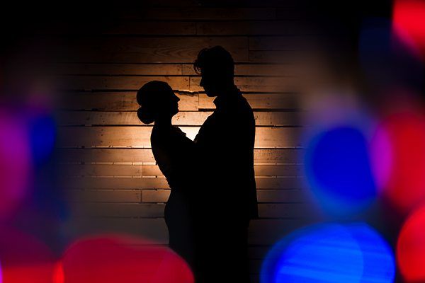 Silhouettes of a couple standing close together against a wooden wall, with blurred colorful lights in the foreground creating a bokeh effect. The lighting creates a soft and romantic ambiance. Image by Documentary Wedding Photographer Lewis Bishop Photography.