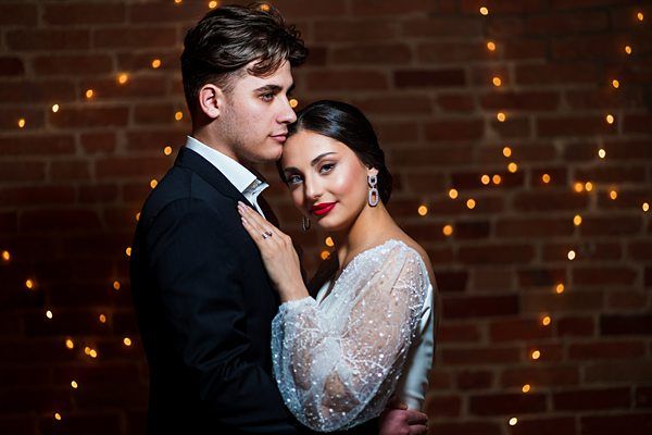 A couple poses together in formal attire against a backdrop of brick and string lights. The man wears a black suit, and the woman wears a white dress with sheer sleeves. The woman rests her hand on the man's chest as they look slightly away from the camera. Image by Documentary Wedding Photographer Lewis Bishop Photography.