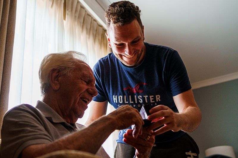 A smiling younger man in a blue Hollister t-shirt shows something on a smartphone to an elderly man with gray hair, who is laughing. They are indoors near a window with beige curtains, and both appear to be enjoying a lighthearted moment together. Image by Documentary Wedding Photographer Lewis Bishop Photography.