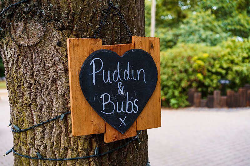 A heart-shaped blackboard sign attached to a tree reads “Puddin & Bubs” in white handwritten text. The tree is wrapped in string lights, and there is lush greenery and a paved area in the background. Image by Documentary Wedding Photographer Lewis Bishop Photography.