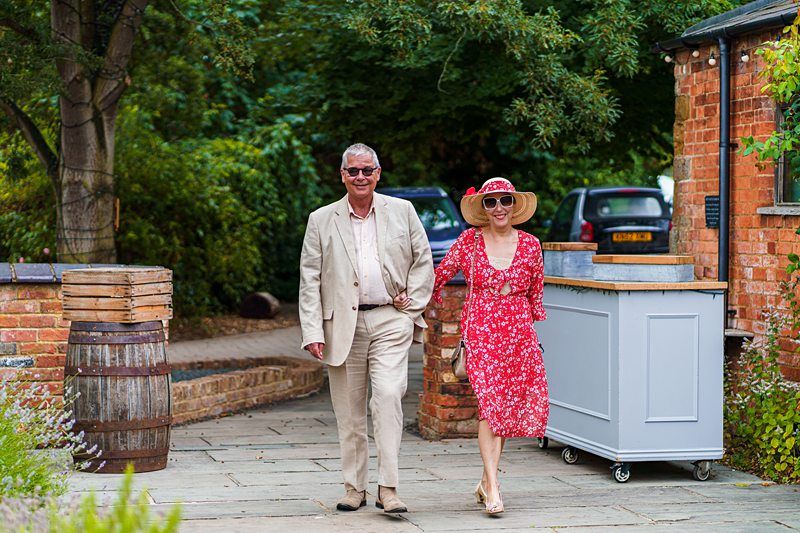 A man in a beige suit and sunglasses and a woman in a red floral dress with a sun hat are walking outdoors on a stone path. They are smiling and enjoying a sunny day, with trees and brick buildings in the background. Image by Documentary Wedding Photographer Lewis Bishop Photography.