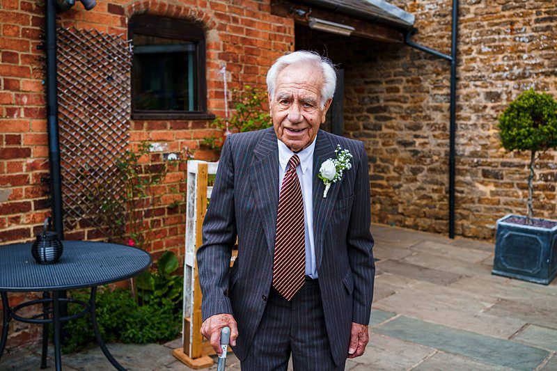 An elderly man with white hair, dressed in a striped suit, white shirt, and patterned tie, stands outside a brick building. He is holding a cane and has a white boutonnière pinned to his lapel. A round table and a potted tree are visible in the background. Image by Documentary Wedding Photographer Lewis Bishop Photography.