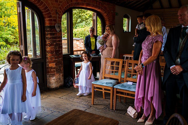 A wedding scene inside a rustic wooden hall. Young flower girls in white dresses stand near a doorway. Guests in formal attire are seated and standing, watching the ceremony. A mother holds a baby while another child stands nearby. The venue has large arched windows. Image by Documentary Wedding Photographer Lewis Bishop Photography.