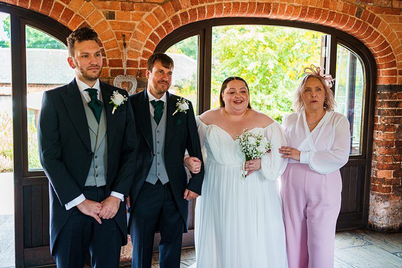 A bride and groom stand together, smiling, with the bride holding a bouquet. They are flanked by two people, possibly family or friends, dressed in formal attire. The group is posed in front of a brick archway with a window displaying a sunny outdoor scene. Image by Documentary Wedding Photographer Lewis Bishop Photography.