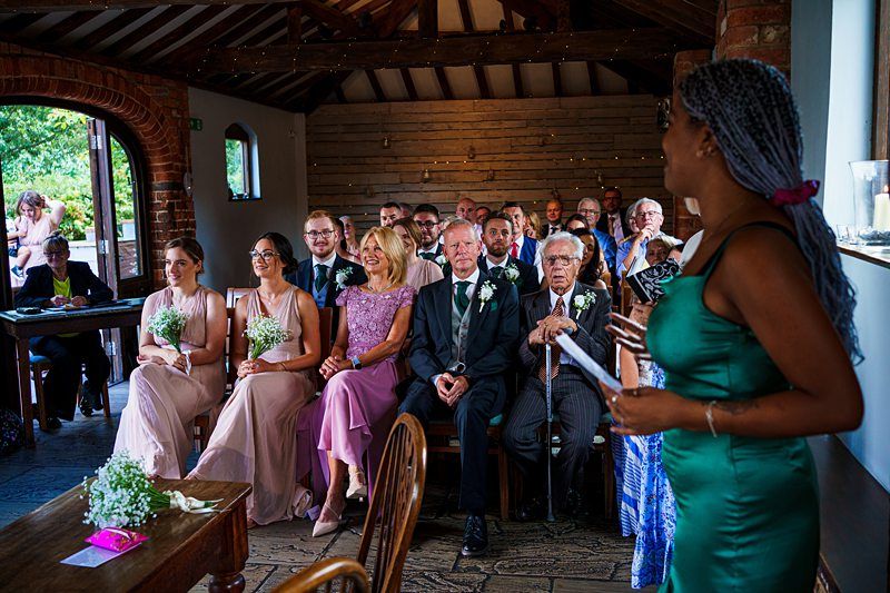 A woman in a green dress is giving a speech to a room of seated wedding guests. The guests include men and women dressed in formal attire, with some women holding bouquets. The setting appears to be a rustic indoor venue with exposed wooden beams and brick walls. Image by Documentary Wedding Photographer Lewis Bishop Photography.