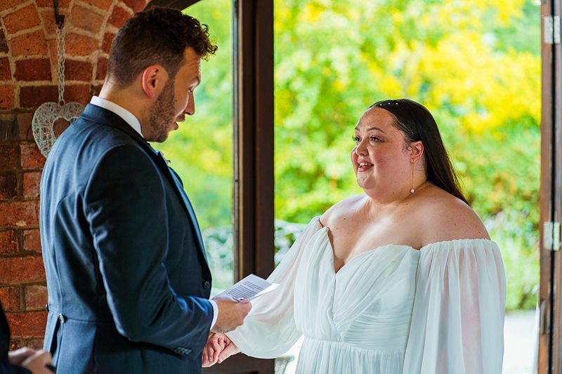 A bride in a white off-the-shoulder dress and a groom in a dark suit are standing indoors near a doorway, holding hands. The bride is smiling and looking at the groom who appears to be reading from a piece of paper. Bright greenery is visible outside. Image by Documentary Wedding Photographer Lewis Bishop Photography.