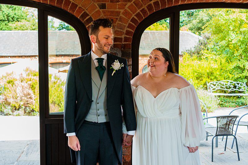 A bride and groom smile and hold hands, standing under an archway with a glass door. The groom wears a dark suit with a white boutonniere, and the bride wears an off-the-shoulder white wedding gown. The background shows a courtyard with greenery and outdoor furniture. Image by Documentary Wedding Photographer Lewis Bishop Photography.
