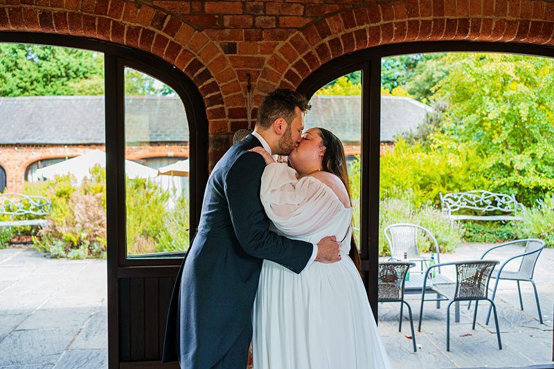 A couple dressed in wedding attire share a kiss under an arched brick structure. The bride wears a white gown, and the groom wears a dark suit. Behind them is a patio with chairs and lush greenery. The moment is full of joy and love. Image by Documentary Wedding Photographer Lewis Bishop Photography.