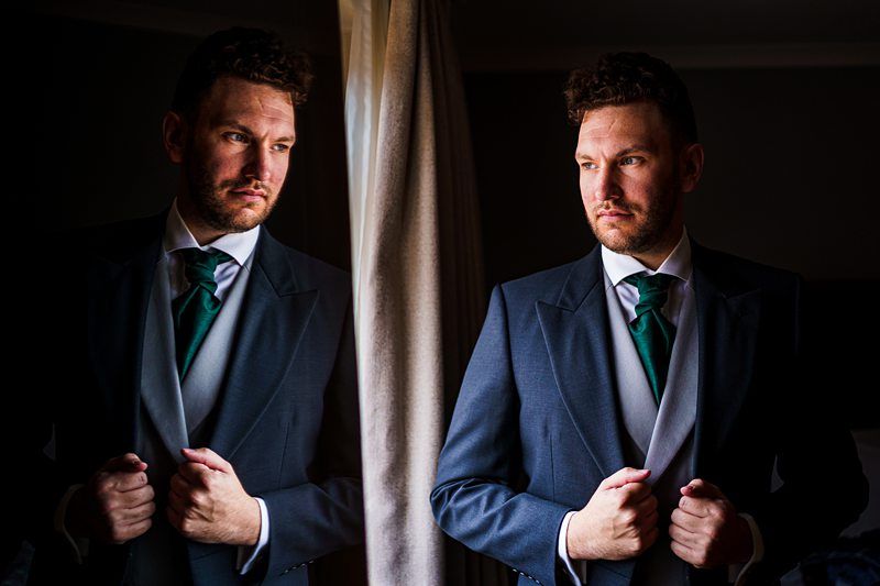 A man in a dark suit and green tie stands facing a mirror, adjusting his jacket. His reflection is clearly visible, mimicking his posture and expression. The background is dimly lit, drawing focus to the man and his reflection. Image by Documentary Wedding Photographer Lewis Bishop Photography.